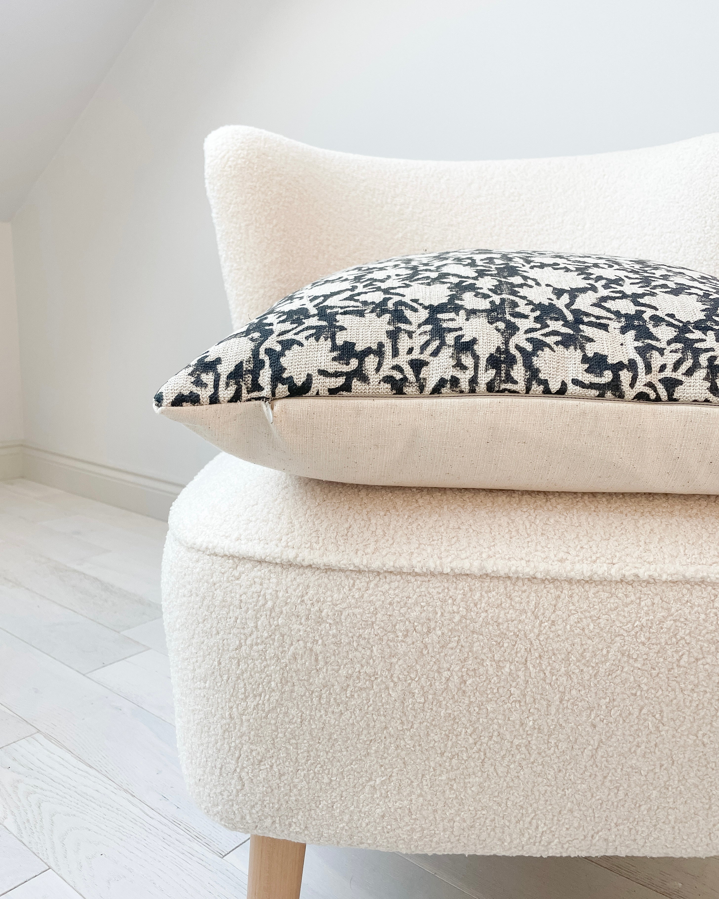 Side view of a 20x20” square luxury heavy linen hand-blocked cushion cover featuring a delicate organic black floral pattern. It has a soft cotton back and an invisible zipper. Hand-block material is produced in Jaipur, India. Cushion cover is made in the UK. Displayed on a neutral cream boucle chair