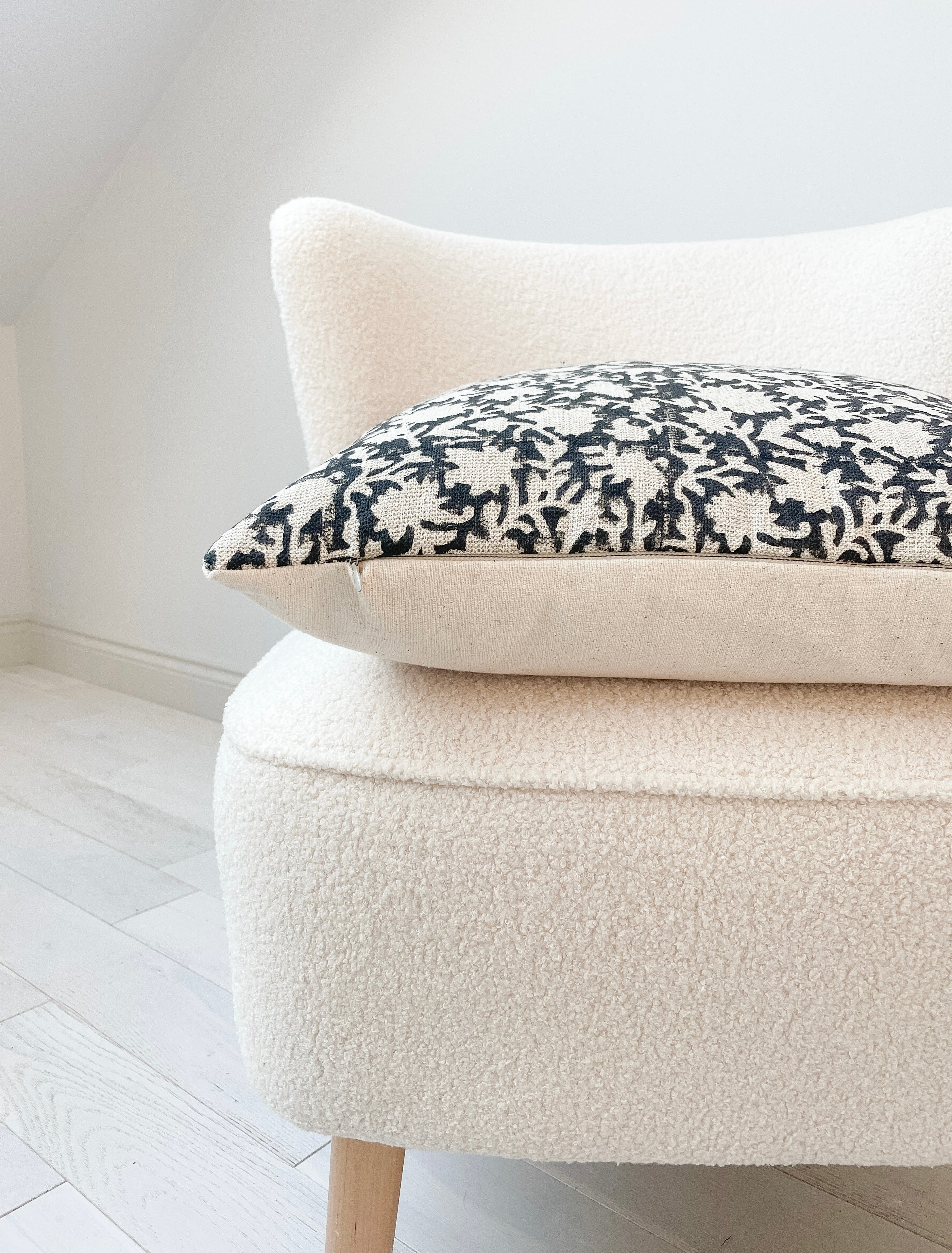 Side view of a 20x20” square luxury heavy linen hand-blocked cushion cover featuring a delicate organic black floral pattern. It has a soft cotton back and an invisible zipper. Hand-block material is produced in Jaipur, India. Cushion cover is made in the UK. Displayed on a neutral cream boucle chair