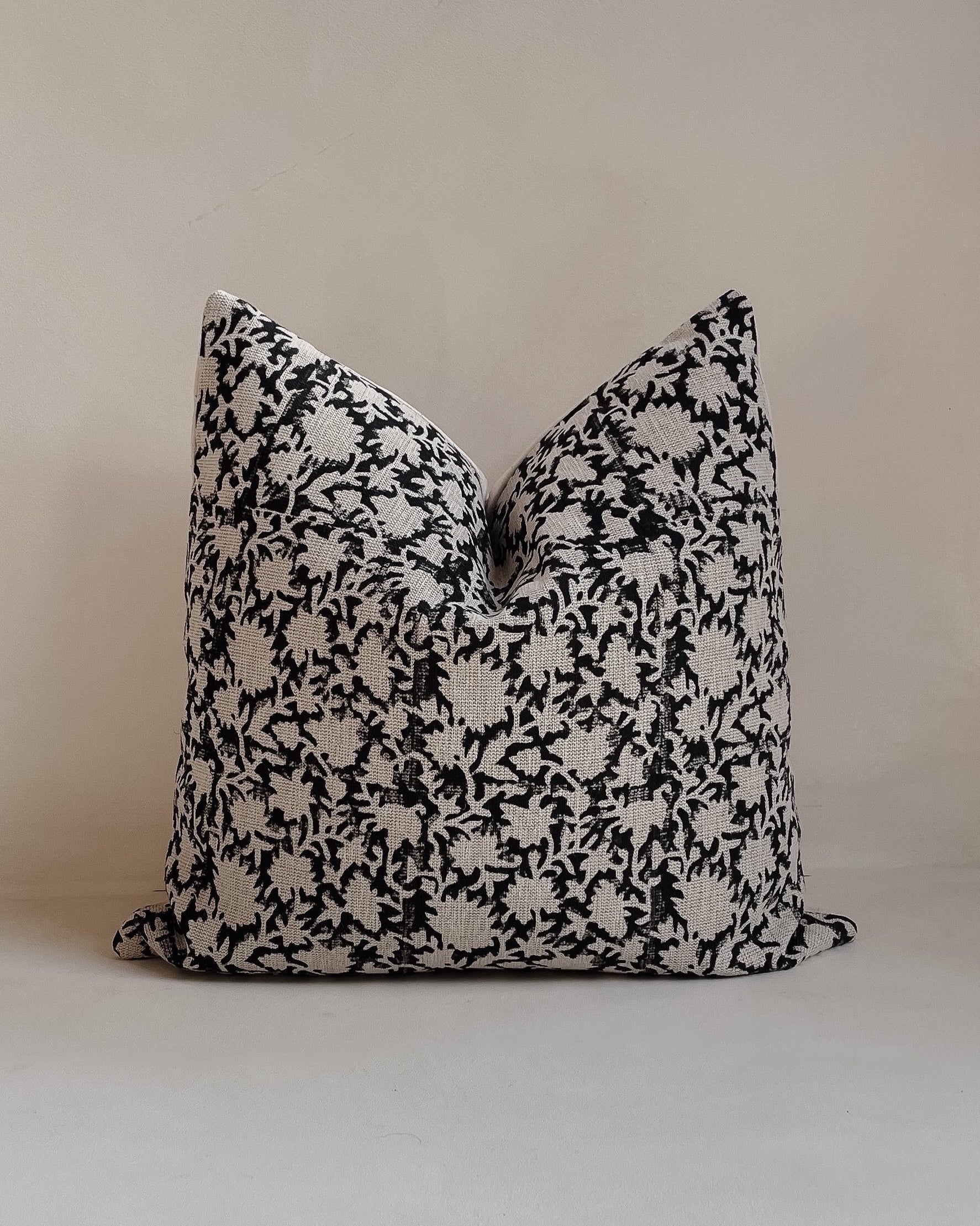 Front view of a 20x20” square luxury heavy linen hand-blocked cushion cover featuring a delicate organic black floral pattern. It has a soft cotton back and an invisible zipper. Hand-block material is produced in Jaipur, India. Cushion cover is made in the UK. Displayed against a neutral limewash background