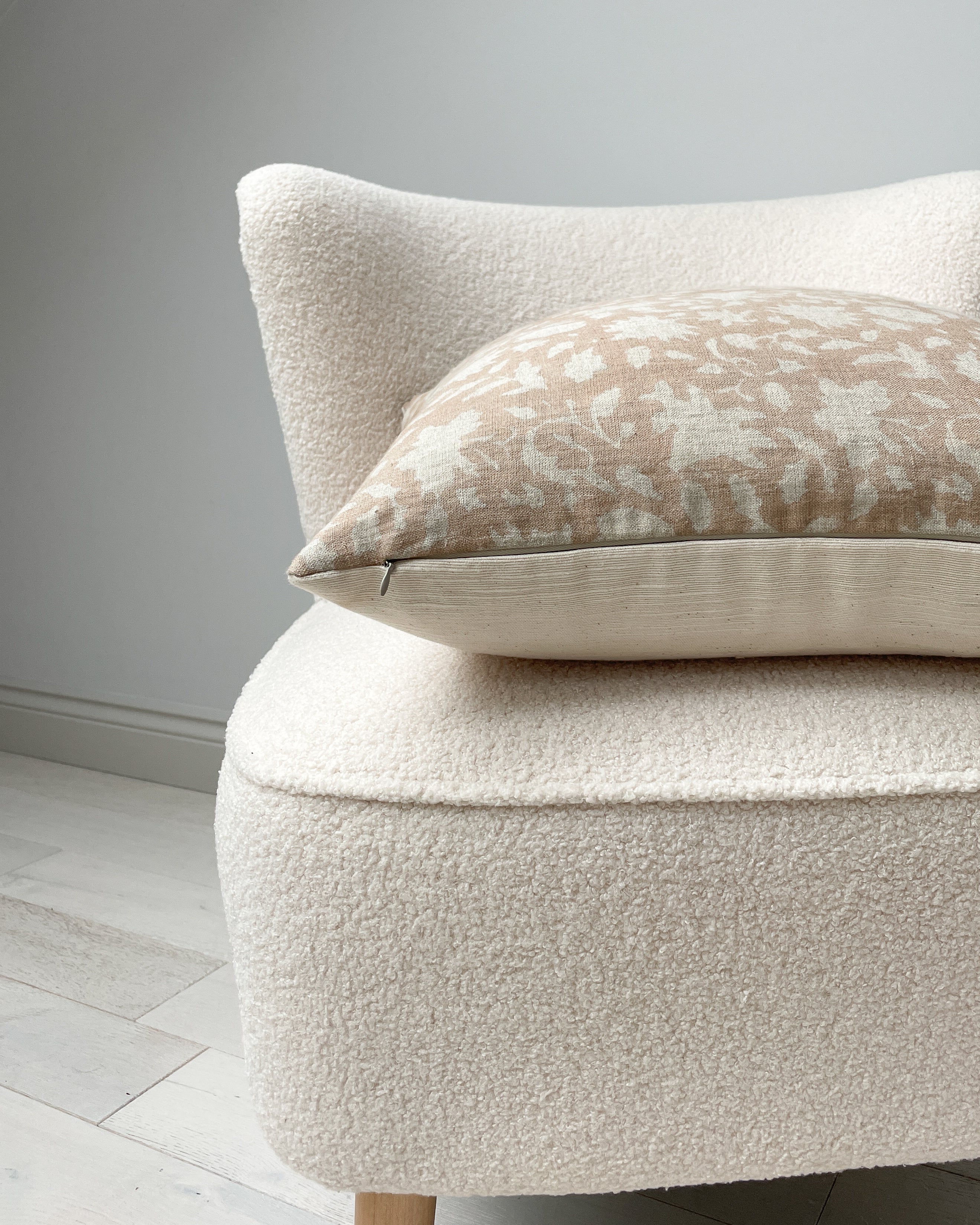 Side view of a 20x20” square luxury natural cotton hand-blocked cushion cover in tan neutral tones featuring a soft muted floral pattern design. It has a textured soft cotton back and an invisible zipper. Hand-block material is produced in Jaipur, India. Cushion cover is made in the UK. Displayed on a cream neutral boucle chair
