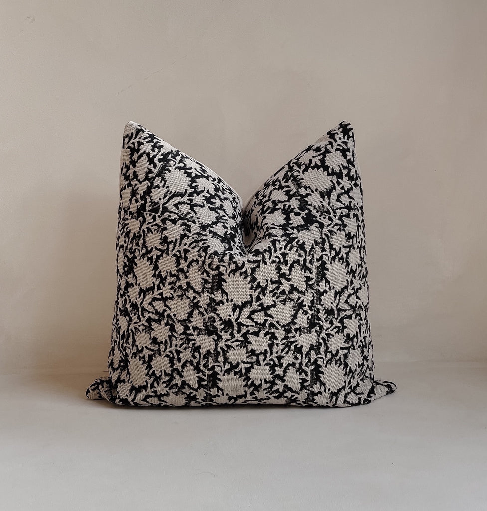 Front view of a 20x20” square luxury heavy linen hand-blocked cushion cover featuring a delicate organic black floral pattern. It has a soft cotton back and an invisible zipper. Hand-block material is produced in Jaipur, India. Cushion cover is made in the UK. Displayed against a neutral limewash background